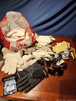 Large bag filled with numerous work gloves, additional gloves and three safety glasses spread on table