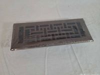 Front view of one vent cover sealed in plastic, showing decorative cutout pattern and brushed copper finish
