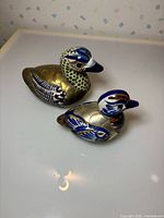 Oblique view of both duck figurines showing brass bodies and painted ceramic sections