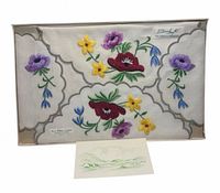Boxed set showing embroidered linen napkin with multicolour flowers and grey scalloped border; small insert card visible
