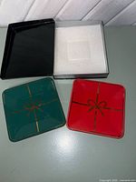 Two square Christmas plates in red and green with gold ribbon motif next to black box and foam insert