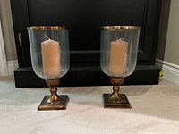Two matching brass-tone base hurricane candle holders with beige pillar candles displayed side by side on carpet