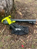 Leaf vacuum/blower lying on ground showing yellow-green motor housing, black tube, wheels, and debris bag