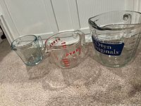 Three glass measuring cups of different sizes and brands on carpeted floor