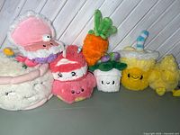 Group of eight Squishable plush toys lined up: clam, macaron, two strawberry cake cubes, mochi, carrot, smoothie cup, chick