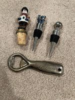 All three wine stoppers and metal bottle opener displayed on carpet
