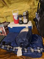 Overall lot showing cooler, lantern, thermoses, beverage jug, Coleman bagged sleeping bag and plaid sleeping bag