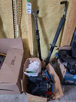 Two trailer jack assemblies leaning against wall beside box of assorted fittings