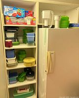 Cabinet next to refrigerator showing stacked plastic containers, pitchers, and boxes on top