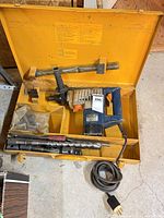 Open yellow metal case showing drill, bits, handle, power cord
