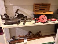 Entire lot on shelves showing combination plane, two hand drills, chisel set, ice skates, and rifle stock