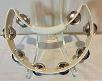 Front view of cream half-moon tambourine on clear stand, showing single row of metal jingles and molded grip