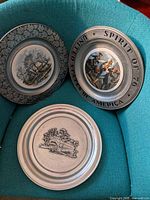 Front view of all three decorative pewter-rim plates showing differing borders and ceramic scenes