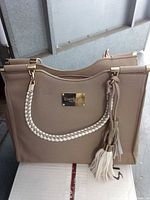 Front view of taupe Bebe handbag showing braided strap, tassels and gold logo plate