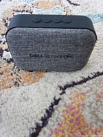 Front view of grey fabric-front Dell Technologies Bluetooth speaker with top control buttons visible