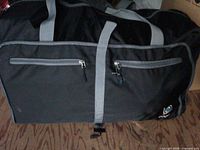 Front view of black Protege duffle bag showing logo, front zip pockets, handles, grey trim