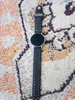 Full view of black round-face smartwatch lying flat, showing ventilated strap and circular display