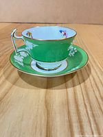Teacup on saucer showing exterior green, floral silhouette décor, gold trim and handle