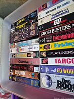 Top view of bin showing two stacks of labeled VHS cassettes including Cats, Ghostbusters, Flatliners, Fargo, Boogie Nights, etc.