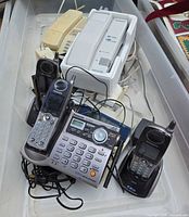 Overview of lot items inside bin: three Panasonic cordless handsets, Panasonic answering base, beige corded phone, chargers, cables