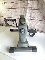 Front view of gray mini pedal exerciser showing pedals, LCD screen and tension dial