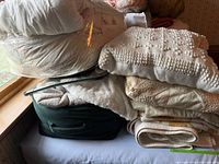 Stacked bedding showing comforter in plastic, chenille bedspread, green zippered bag, folded blankets