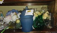 Shelf view showing blue ceramic jar, artificial flowers, green glass vase, small glass dish, and portion of figurine