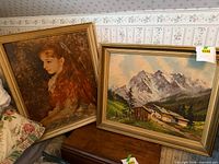Both framed artworks leaning against wall: mountain landscape oil and Renoir portrait print