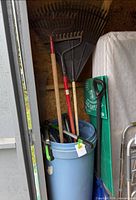 Trash can with visible rakes, shovel handles, and surrounding items