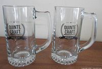 Front view of two Quinte Craft glass mugs showing logo and size