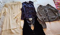 Four clothing pieces laid out: off-white suit, black velvet vest, purple satin vest, grey sweater