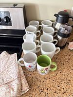 Countertop with ten white mugs, two novelty mugs, stainless steel carafe, coffee grinder, floral textile