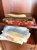 Stack of brown runner, burgundy floral cloth, white/blue and white/orange linens on shelf