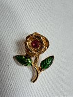 Front view of gold-tone rose pin showing green enamel leaves and red center stone