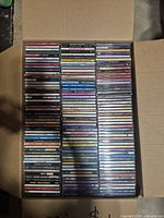 Open cardboard box filled with two tightly packed rows of audio CDs in jewel cases, spines visible