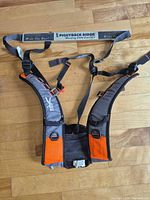 Front view of Piggyback Rider harness showing full unit with straps, buckles, orange/gray panels and attached metal foot bar