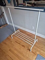 Full view of white metal garment rack showing top rail, adjustable side posts, lower shoe shelf and caster wheels
