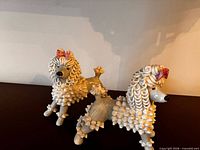 Two small ceramic poodle figurines with textured fur, pink bows, displayed side by side