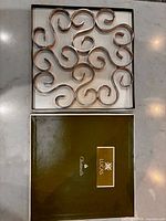 Top view of swirl-pattern metal trivet in open box with branded lid