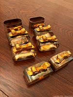 Eight rectangular silver-tone napkin rings each topped with a gold-tone bow displayed on wood surface