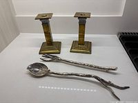 Two brass-coloured candle holders and two branch-form salad tongs displayed together