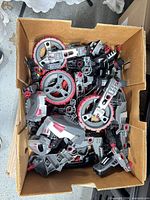 Box of assorted gray, red and black plastic construction toy pieces including large wheel discs and various connectors
