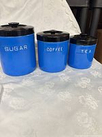 Set of three blue metal canisters labeled Sugar, Coffee, Tea with black lids