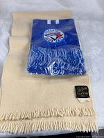 Beige Glen Currie scarf with packaged blue Blue Jays scarf on top