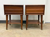 Front view of both matching nightstands showing drawers, lower shelves, tapered legs, brass caps