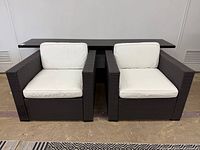 Front view of two matching dark brown resin wicker lounge chairs with white cushions