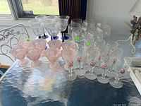 Four complete glassware sets displayed on table: blue stem glasses, pink sherbet cups, floral goblets, etched snifters