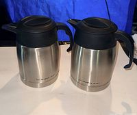 Two identical stainless steel insulated pitchers with black lids and handles side by side
