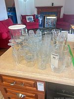 Group view of glassware lot on countertop showing trifle bowl with lid, stemmed glasses, tumblers, assorted small glasses, cake stand, large vase-style glass