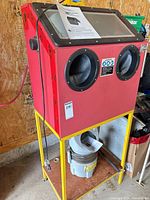 Red blast cabinet on yellow stand with hopper beneath and manual on window